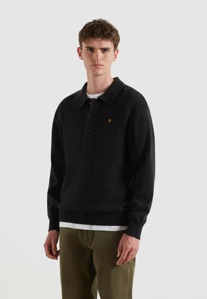 Maglione polo nero a maglia con colletto, con bottoni sul davanti e un logo arancione ricamato. Indossato sopra una t-shirt bianca e abbinato a pantaloni verdi.