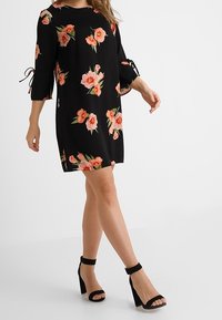Vestido negro con flores, de manga tres cuartos, con flores rosas y naranjas, y un corte holgado. Lleva consigo unos tacones negros con tira en el tobillo.