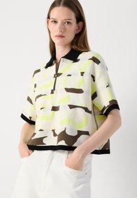 Cropped polo shirt featuring a black collar, zip front, and a camouflage pattern in shades of cream, brown, and neon green. Short sleeves.