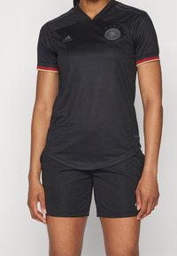 Maglia atletica nera con maniche corte, scollatura a V e una sottile texture a rete. Presenta dettagli rossi e dorati sulle maniche e un logo con stemma. Pantaloni neri.