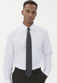 Balta ilgomis rankovėmis mygtukinė marška su lygiu paviršiumi, derinama su tamsiai pilku dryžuotu kaklaraiščiu. Marška įkišta į tamsius kelnes.