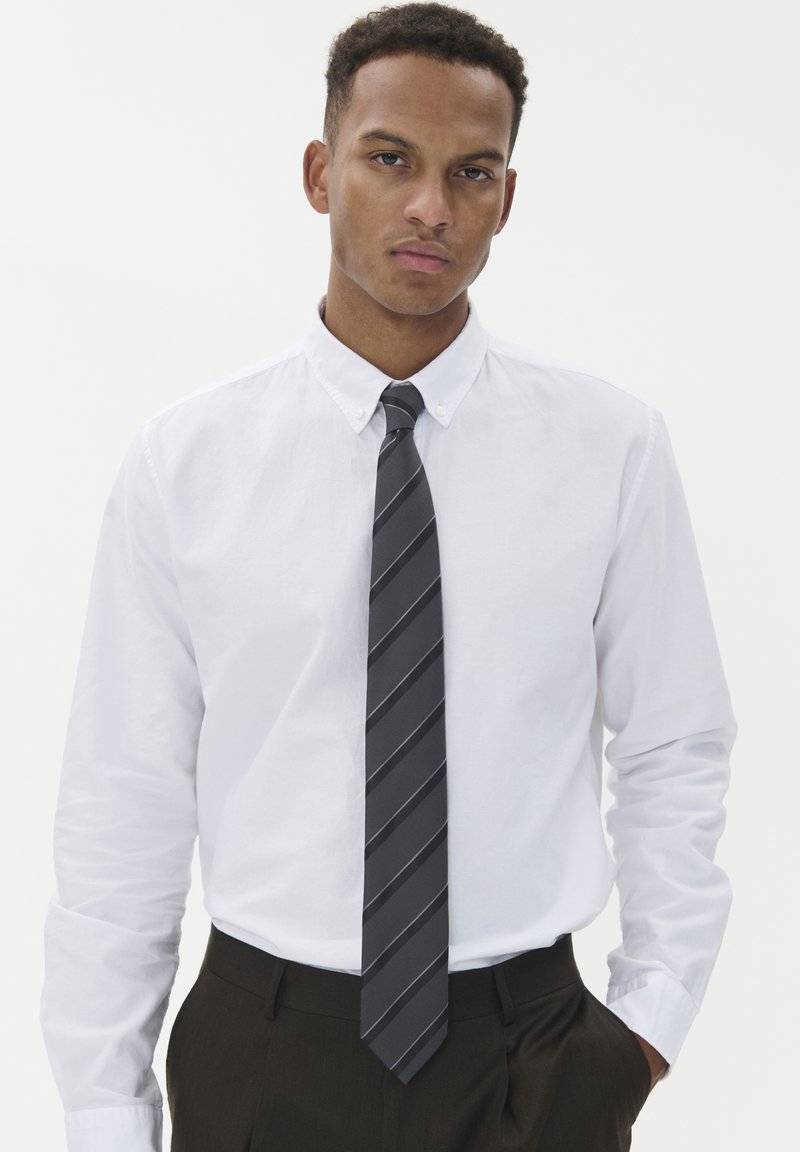 Balta ilgomis rankovėmis mygtukinė marška su lygiu paviršiumi, derinama su tamsiai pilku dryžuotu kaklaraiščiu. Marška įkišta į tamsius kelnes.