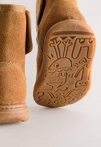 Ghete scurte din piele întoarsă de culoare cacao, având o talpă din cauciuc texturată cu gravuri jucăușe și detalii cusute de-a lungul marginilor.