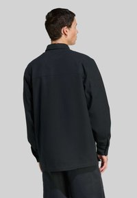 Muž s krátkymi tmavými vlasmi, oblečený v voľnej čiernej košeli s dlhým rukávom a čiernych nohaviciach, stojí otočený chrbtom proti jednoduchému šedému pozadiu.