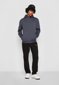 Felpa grigia con colletto alto, abbinata a pantaloni sportivi neri. L'outfit è completato da scarpe bianche e un berretto nero con logo.