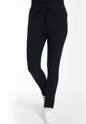 Zwarte joggingsbroek van zacht materiaal, met een koordtaille, slim fit en schuine zijzakken. G gedragen met witte sneakers.