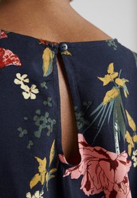 Dettaglio ravvicinato della scollatura posteriore di un abito floreale blu scuro con chiusura a bottone e apertura a occhio di bue, che mostra la pelle sottostante.