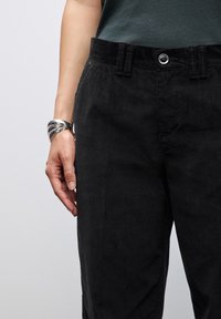 Pantaloni in velluto a coste neri con taglio dritto, dotati di chiusura frontale con bottone e passanti per cintura, abbinati a un bracciale in argento su una mano.