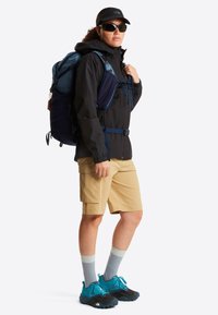 Άτομο που φορά μαύρο μπουφάν, ταμπά cargo σορτς, μπλε παπούτσια πεζοπορίας, γυαλιά ηλίου και σακίδιο πλάτης, στέκεται στραμμένο προς τα αριστερά σε λευκό φόντο.