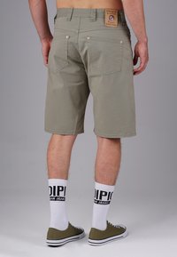 Grüne Baumwollshorts mit geradem Schnitt, fünf Taschen, dezenten Nähten und einem hellbraunen Markenetikett am hinteren Bund. Weiße Socken mit schwarzem Text.