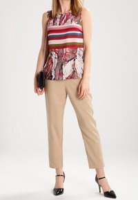 Kobieta w bezrękawnikowej, wielokolorowej w paski bluzce, beżowych, krótkich spodniach, czarnych, spiczastych szpilkach, trzymająca małą czarną torebkę na ramieniu.