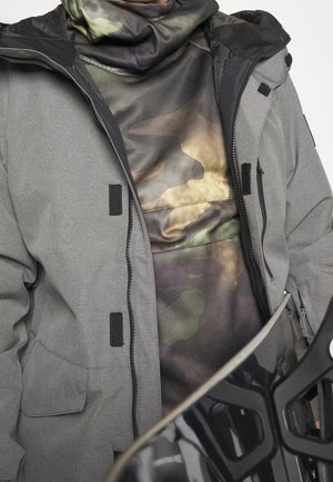 Person iført en grå jakke over en camouflage hættetrøje, der holder et sort snowboard med synlige bindinger.