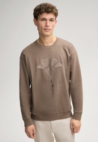 Felpa marrone in cotone con un design testurizzato ricamato che presenta una forma floreale stilizzata. Collo a giro e polsini a costine.