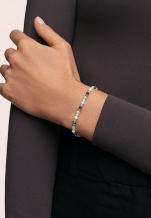 Pulsera de cuentas con acentos en azul claro, verde y dorado. Cuentas lisas y redondas de varios tamaños, llevada en una muñeca contra una tela oscura.