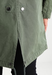 Veste vert olive à fermeture à bouton pression, fentes latérales et cordon de serrage ajustable à l'ourlet. Fabriquée en tissu léger et texturé.