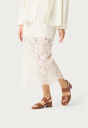 Mujer con falda midi de encaje blanca, blusa color crema y sandalias marrones de tacón bajo caminando frente a un fondo liso y claro.