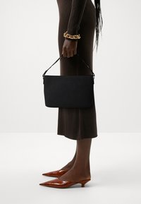 Bolso de mano rectangular negro con un perfil delgado y un asa corta, combinado con un vestido marrón acanalado y tacones marrones de punta. Pulsera de cadena dorada visible.