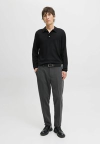 Camisa de polo negra de manga larga con cuello, combinada con pantalones gris claro con pliegues y zapatos negros de cordones, sobre un fondo blanco liso.