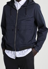 Navy zip-up jacket with a hood, featuring two front pockets, a smooth texture, and elastic cuffs. Layered with a patterned shirt underneath.