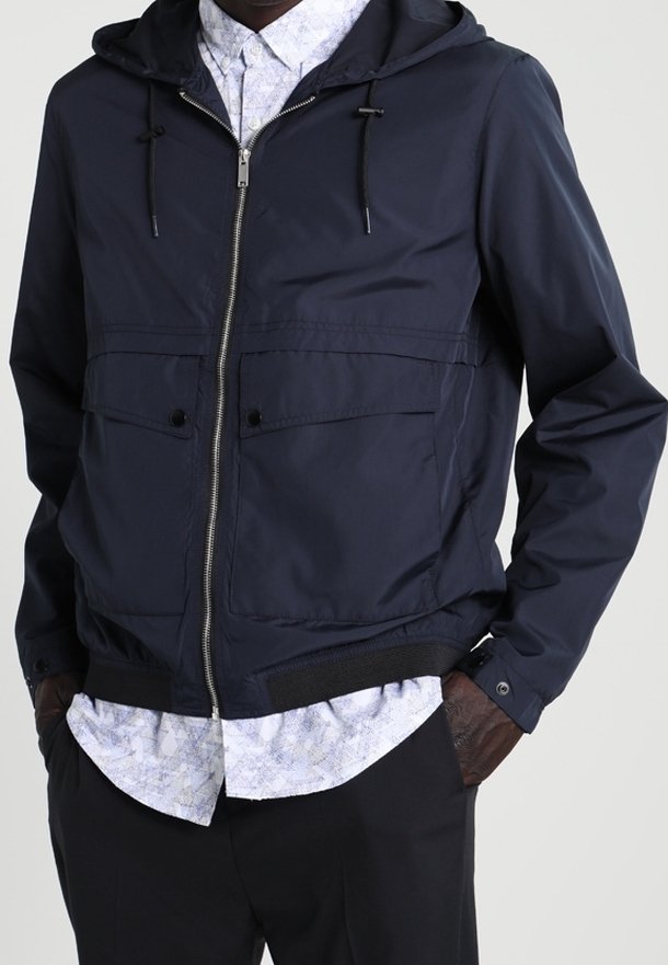 Navy zip-up jacket with a hood, featuring two front pockets, a smooth texture, and elastic cuffs. Layered with a patterned shirt underneath.
