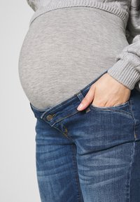 Jeans de gravidez em tecido de denim azul, com uma cintura de altura média e painéis laterais elásticos para conforto. Blusa cinza com textura canelada.