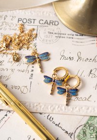 Guldhalsband och örhängen med blå trollsländehängen liggande på vintage handskrivna vykort bredvid en guldfärgad penna och ett mässingsföremål.
