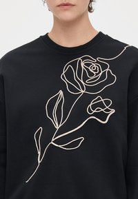 Μαύρη μπλούζα με στρογγυλή λαιμόκοψη και relaxed fit, με σχέδιο λουλουδιού σε γραμμή με μπεζ κλωστή, που απεικονίζει δύο τριαντάφυλλα και φύλλα.