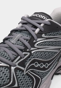 Γκρι και μαύρο αθλητικό παπούτσι με ύφασμα από δίχτυ, γκρι κορδόνια και αντανακλαστικές ασημένιες λεπτομέρειες, εμφανίζοντας το λογότυπο της μάρκας στο πλάι και στη γλώσσα.