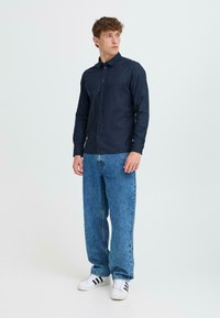 Camisa de manga larga azul oscura con cuello, frente abotonado, combinada con jeans sueltos de un azul claro y zapatillas blancas con rayas negras.