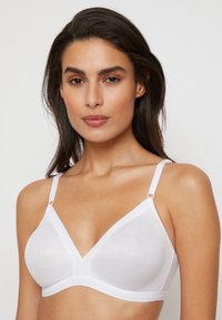Bralette blanc en coton avec une forme triangulaire, texture côtelée, bretelles ajustables et accents métalliques décoratifs sur les connecteurs de bretelles.