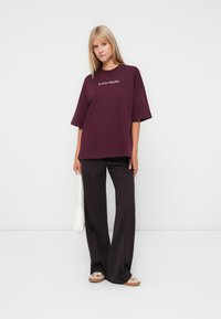 Oversized burgundy t-shirt with short sleeves, featuring white "LeGer Studio" text. Paired with black wide-leg pants and a white handbag.