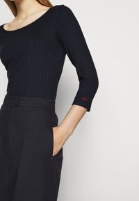 Frau trägt ein schwarzes, geripptes Oberteil mit 3/4-Ärmeln und hochgeschnittene schwarze Hosen, Hand in die Hosentasche gesteckt, "HUGO" auf dem Ärmel eingefasst.