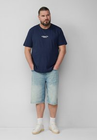 Mężczyzna z brodą, ubrany w granatową koszulkę s.Oliver Co., jasne dżinsowe szorty, białe skarpetki i białe trampki, stoi z rękami w kieszeniach.