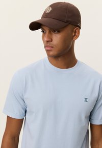 Γαλάζιο μπλουζάκι με γυρισμένα μανίκια, με μικρό πράσινο λογότυπο "II". Σκούρο καφέ καπέλο με κέντημα λογότυπου και καμπυλωτό γείσο.