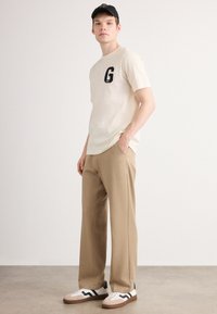 Jeune homme portant un t-shirt beige avec un grand "G", un pantalon beige, des baskets blanches avec des rayures noires, et une casquette de baseball noire, debout à l'intérieur.