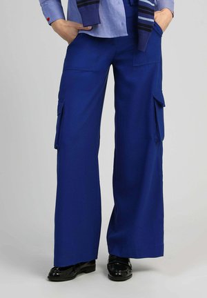 Personne portant un pantalon cargo bleu roi à jambes larges avec des poches avant et latérales, assorti à des chaussures noires brillantes, les mains dans les poches.