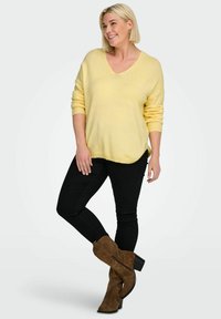 Pull en maille jaune avec un col en V, des poignets côtelés et des fentes latérales, associé à un jean skinny noir et des bottines en daim marron.