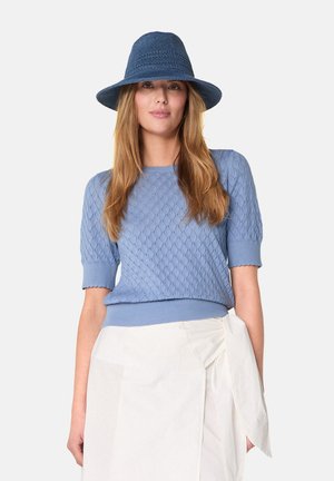  Lichtblauwe textuur gebreide trui met poffende mouwen, in combinatie met een witte wikkelskirt. Een bijpassende strohoed maakt de outfit compleet.