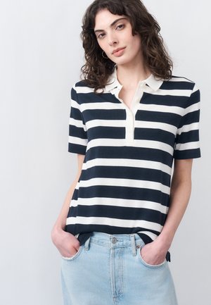 Νεαρή γυναίκα με σγουρά μαλλιά που φοράει μαύρο και άσπρο πόλο μπλουζάκι με οριζόντιες ρίγες, το οποίο είναι μπα tucked μέσα σε ανοιχτόχρωμο τζιν, με τα χέρια στις τσέπες.