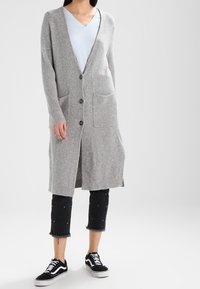 Grå, lång kofta gjord av mjukt stickat material, med V-ringning, knäppning och framfickor. Bärs över en ljusblå topp, ihop med svarta caprijeans.