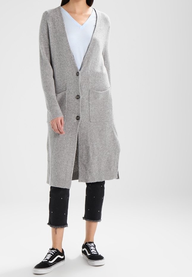 Grå, lång kofta gjord av mjukt stickat material, med V-ringning, knäppning och framfickor. Bärs över en ljusblå topp, ihop med svarta caprijeans.