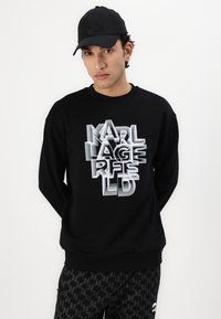 Svart sweatshirt med en framträdande 3D-vit "KARL LAGERFELD"-logotyp. Ribbad halsringning och ärmslut. Matchad med svarta mönstrade byxor.