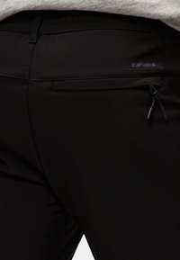 Gros plan sur un pantalon en tissu noir avec une poche à fermeture éclair à l'arrière et une étiquette de marque cousue à la taille.
