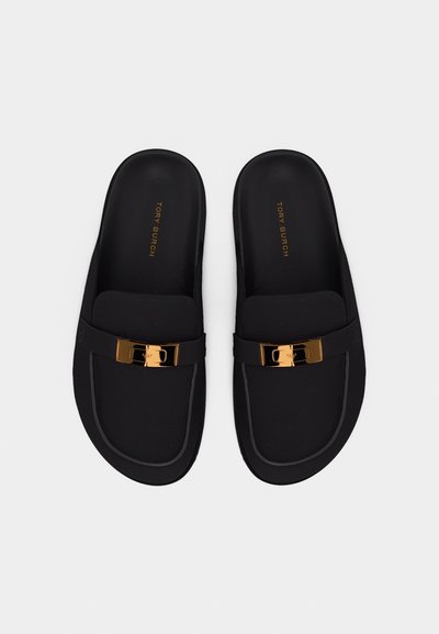 Μαύρα μοκασίνια Tory Burch slip-on με μεταλλικές λεπτομέρειες σε χρυσό τόνο στο πάνω μέρος, απεικονιζόμενα από πάνω επάνω σε ανοιχτό φόντο.
