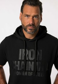Černá mikina s kapucí, která má velký, vyvýšený černý nápis "IRON CHAIN IND. POWER OF STEEL." Hladká tkanina s volným střihem.