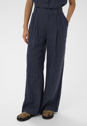 Sötétkék bő szárú nadrág fehér, vékony csíkokkal, barna fonott szandállal viselve, kezek lazán a zsebekben, egyszerű háttér előtt.