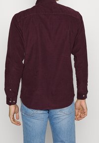 Man som bär en långärmad mörklila kord skjorta och ljusblå jeans, sedd bakifrån mot en enkel bakgrund.