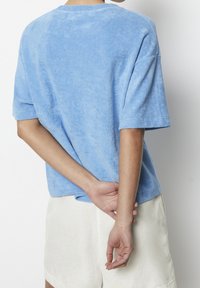 Ljushblå, kortärmad t-shirt i frottétyg med avslappnad passform och rund halsringning, sedd bakifrån med händerna sammanflätade bakom ryggen.