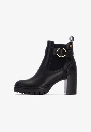 Botín negro de cuero hasta el tobillo con un tacón grueso, paneles laterales elásticos, detalle de hebilla circular y suela texturizada. Cuenta con una pestaña en la parte trasera.