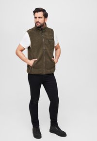 Fleeceweste in dunkelolivgrün mit hohem Kragen, Vorderreißverschluss und Seitentaschen. Kombiniert mit einem weißen T-Shirt und schwarzen Jeans.